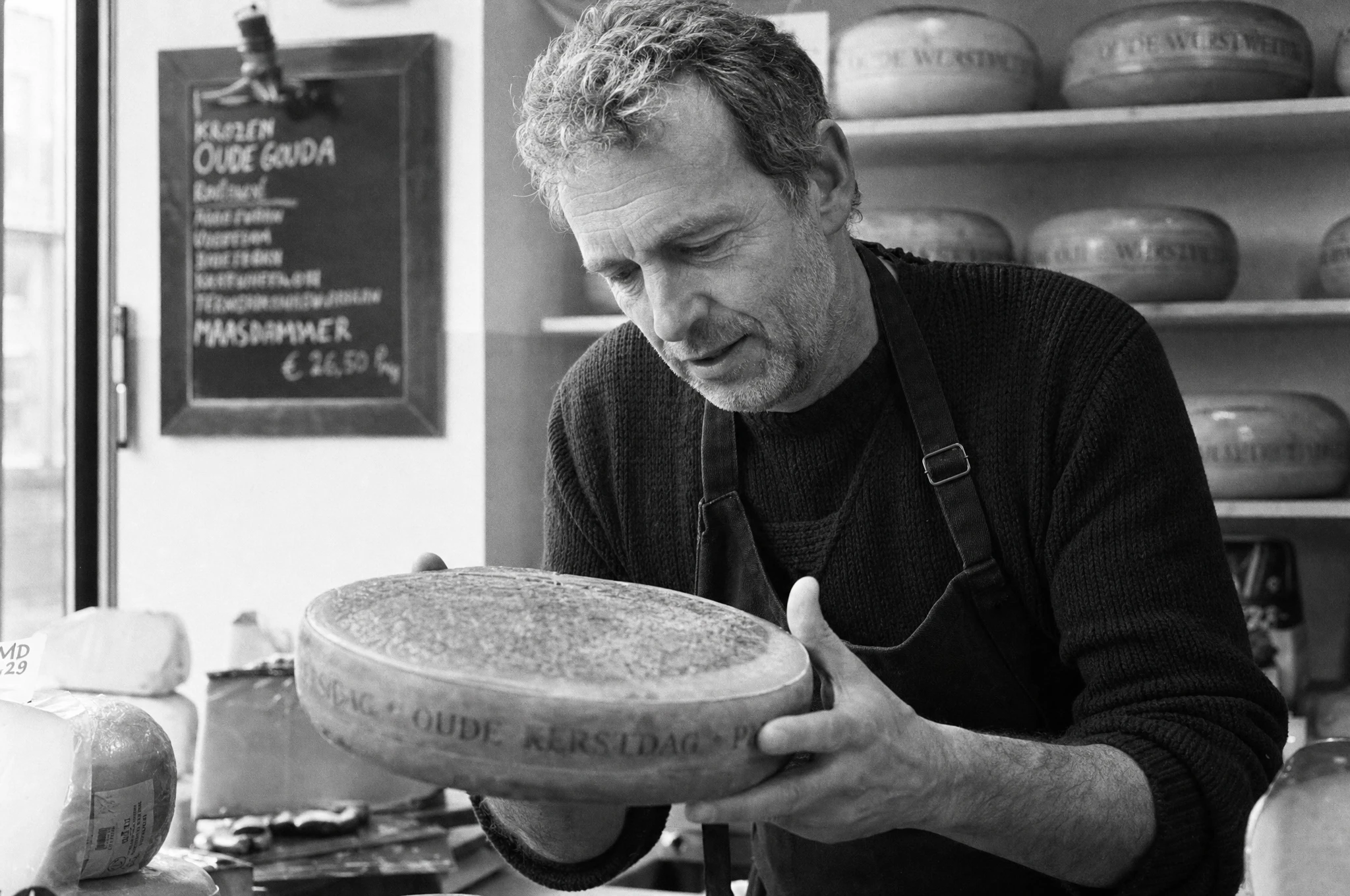 Middle-aged man in his 40s examining a wheel of aged gouda at a European cheese shop counter, documentary black-and-white photograph, Leica M6 with 50mm f/2 Summicron, Ilford HP5 Plus 400, soft window light from the left, visible stubble and weathered hands, unretouched grain, photojournalistic editorial style